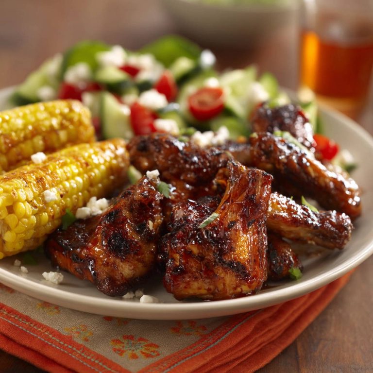Klebrige BBQ Hähnchenflügel vom Grill, nah aufgenommen mit gegrilltem Mais und Salat garniert.