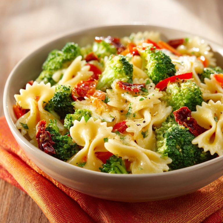 Farfalle mit Brokkoli und getrockneten Tomaten als Nahaufnahme in einer Schüssel auf einem orangen Tuch.
