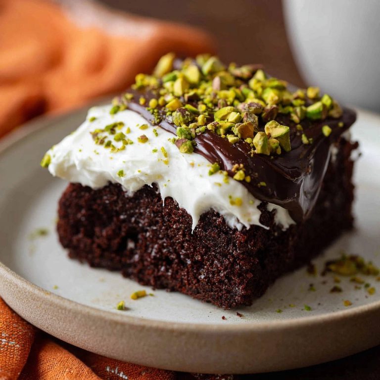 Ein saftiges Stück Schoko-Pistazien-Kuchen mit Sahne und Schokoladenglasur, garniert mit gehackten Pistazien, auf einem Teller serviert.