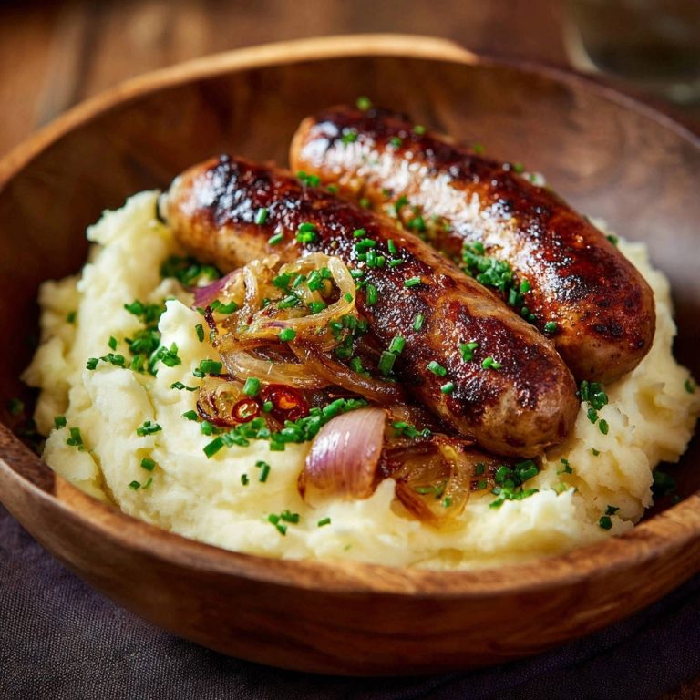 Saftige Bratwürste auf cremigem Püree mit karamellisierten Zwiebeln, Nahaufnahme der Garnitur mit Schnittlauch und Chili.