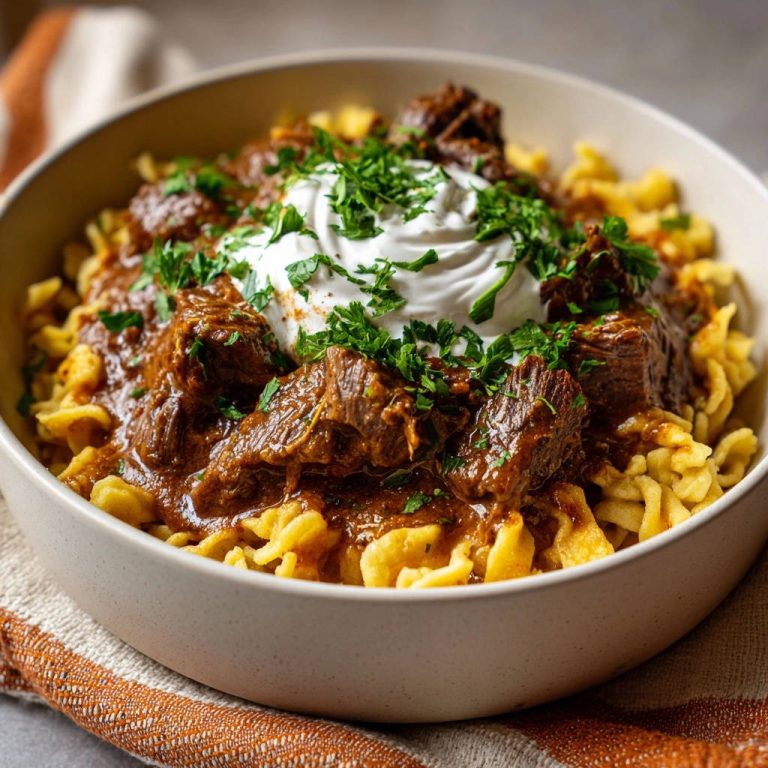 Herzhaftes Rindergulasch mit Spätzle, garniert mit einem Klecks Sauerrahm und frischer Petersilie, in einer Schüssel serviert.